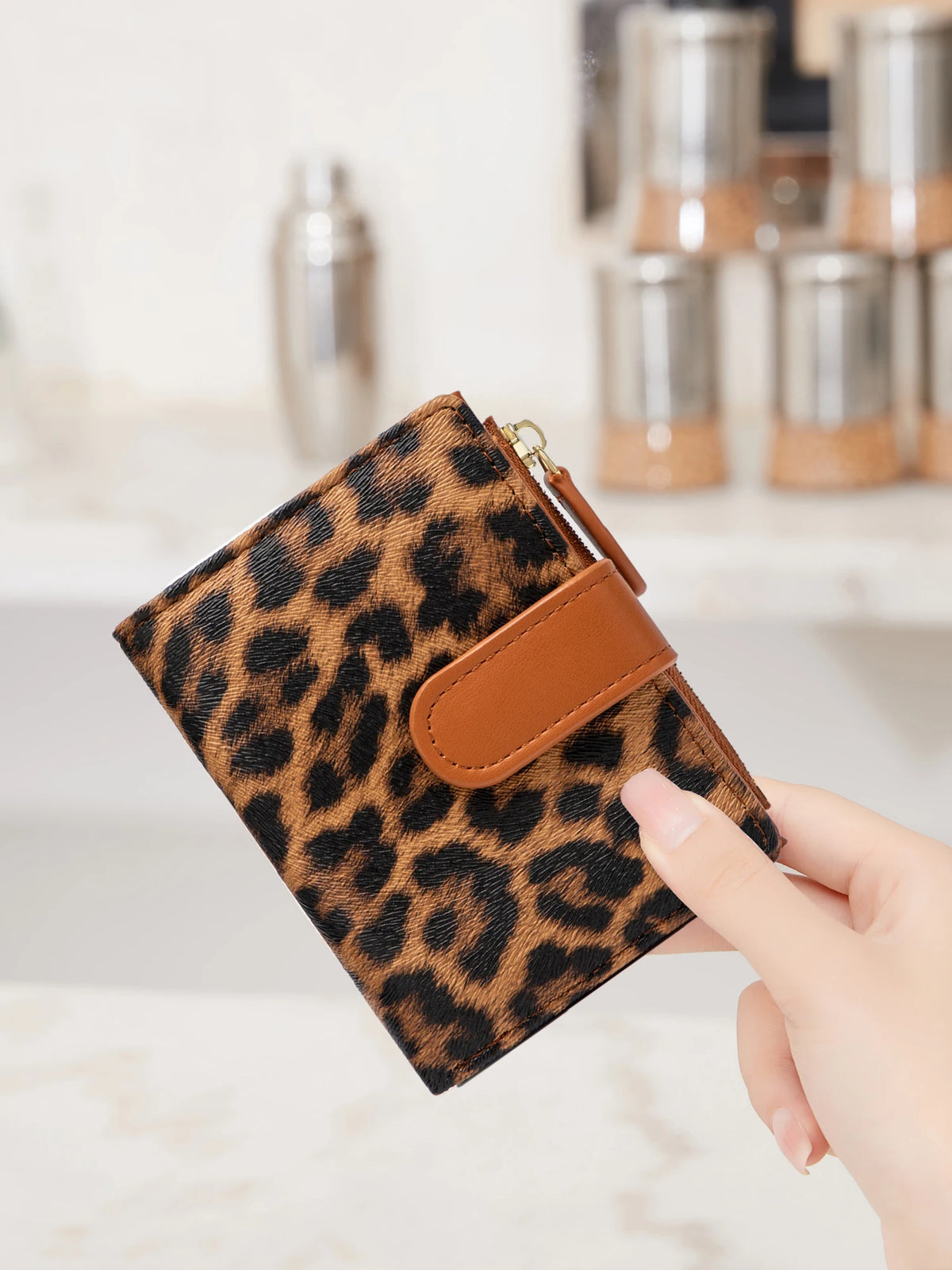A hand holds a brown leopard print compact zippered wallet with a leather strap and card slots.