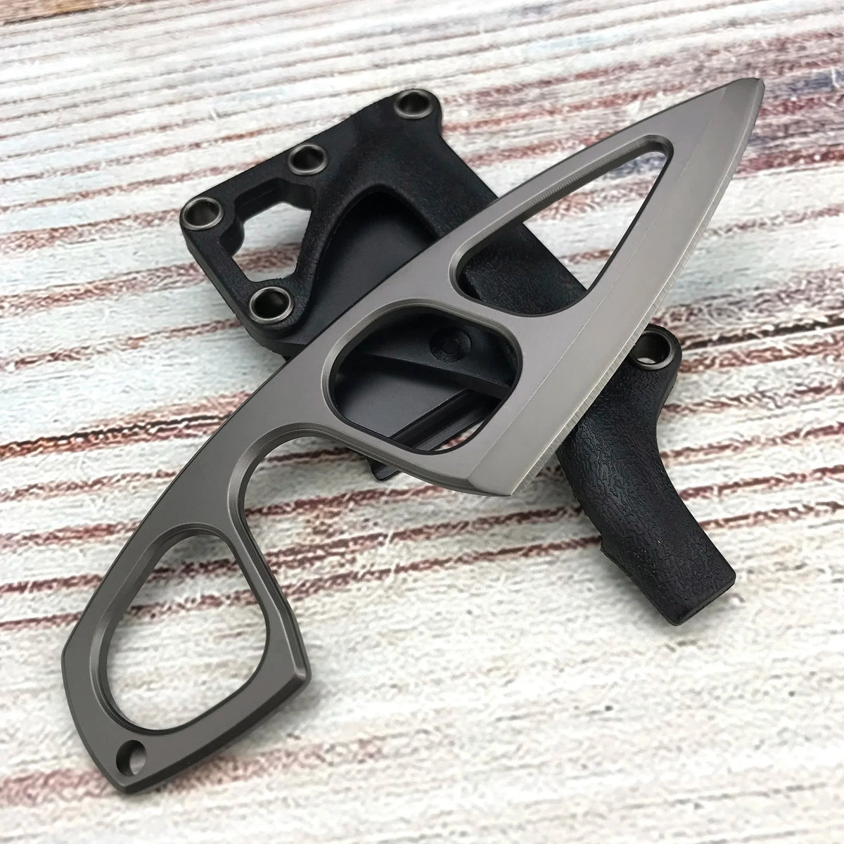 A compact fixed-blade neck knife with a black handle and silver blade is shown on a light wooden surface.