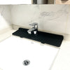 A black silicone faucet splash mat with a metal faucet is placed on a white countertop.