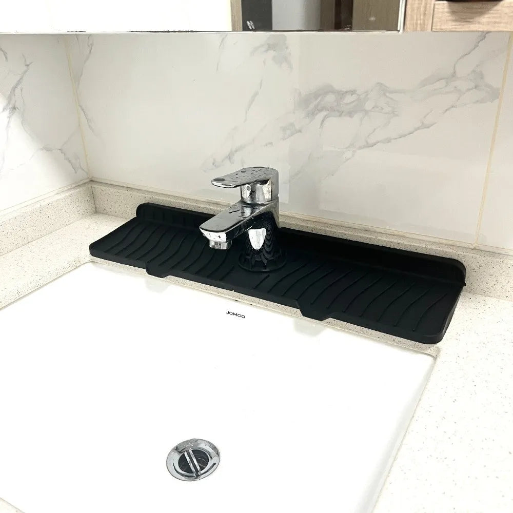 A black silicone faucet splash mat with a metal faucet is placed on a white countertop.