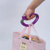 A hand is holding a purple ergonomic grocery bag carrier with a soft-grip handle above a pink striped bag.