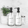 A set of two clear glass hand soap dispensers with black pumps and stainless steel accents on a white surface.
