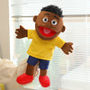 A brown hand puppet with black curly hair, a yellow shirt, blue shorts, and red shoes is shown against a window background.