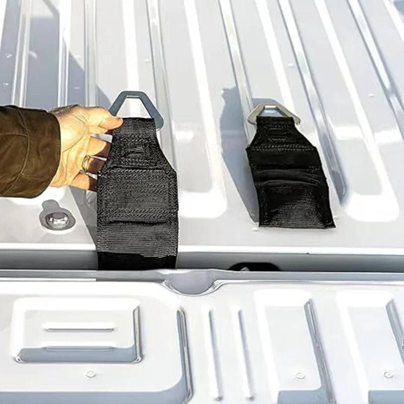 A hand is placing a black fabric bed anchor tie-down into the latch of a truck tailgate.