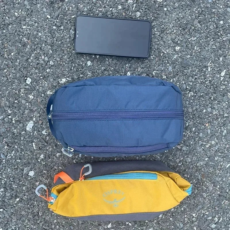 Un sac de toilette pliable et étanche bleu foncé et un smartphone noir posés sur du gravier gris.