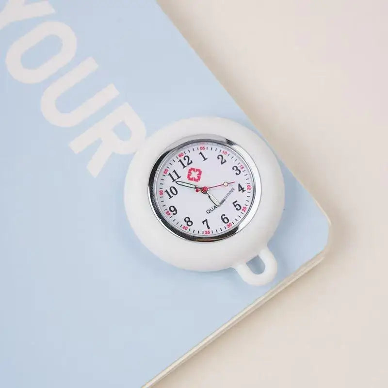 A white silicone pocket watch with a pink and red face is shown resting on a light blue notebook.