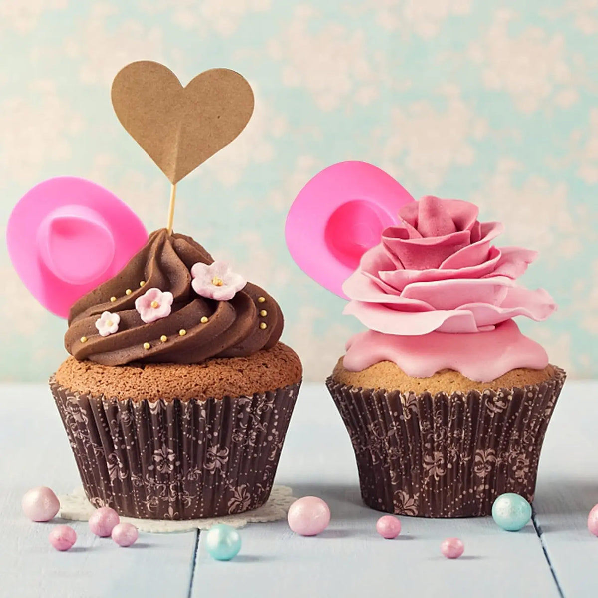 A cupcake decorated with chocolate frosting and topped with a pink heart-shaped paper cutout and a pink rose swirl.