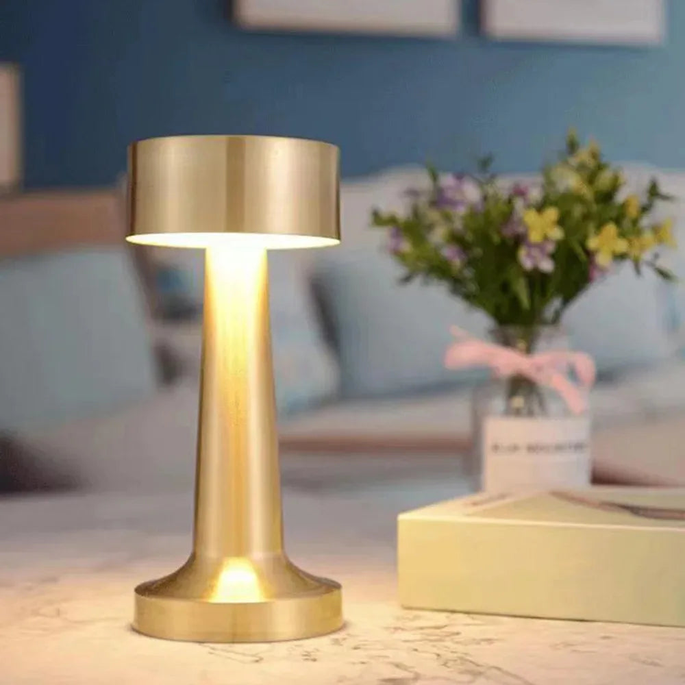 A brass-colored rechargeable metal LED table lamp with a glowing light illuminates a surface.