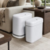 A white, slim rectangular trash can set with lids is displayed on a wooden table next to a sofa.
