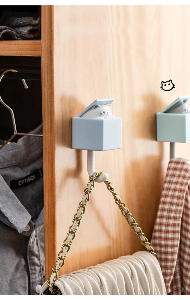 A light blue cat-shaped adhesive hook is mounted on a wooden cabinet door, hanging from a gold chain.