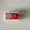 A pink divided container holds a round brown object and three red strawberries, attached to a gold chain.