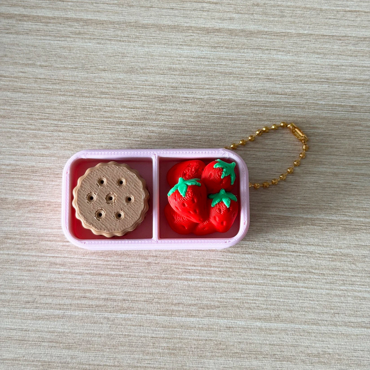 A pink divided container holds a round brown object and three red strawberries, attached to a gold chain.