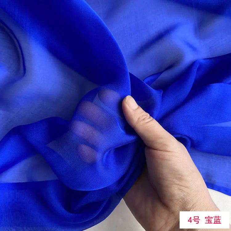 A hand is folding a piece of bright blue, sheer silk chiffon fabric.