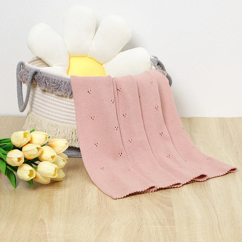 A pink knitted swaddle blanket is draped over a white and yellow flower-shaped storage basket, with yellow tulips nearby.