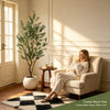 An artificial indoor olive tree in a pot is displayed next to a beige armchair in a sunlit room.