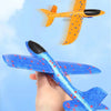 A hand holds an orange foam glider plane toy, with a yellow and black version flying in the background against a light blue s