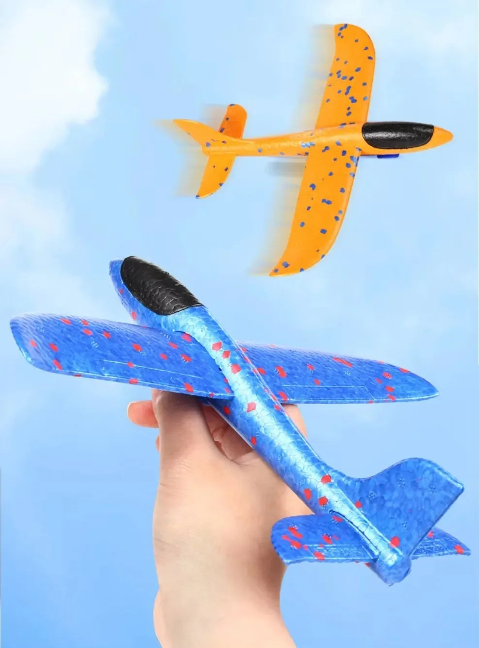 A hand holds an orange foam glider plane toy, with a yellow and black version flying in the background against a light blue s