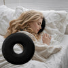 A blonde woman rests her head on a black memory foam ear piercing pillow in a white bed.