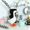 A white goose sculpture with an orange beak and legs is positioned in front of a potted plant and various mugs on a white des
