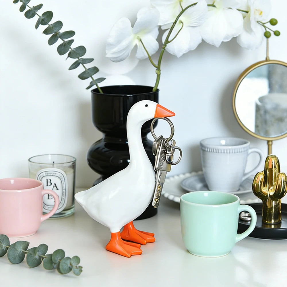 A white goose sculpture with an orange beak and legs is positioned in front of a potted plant and various mugs on a white des