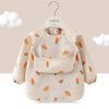 A beige children's waterproof long sleeve feeding bib with a catch pocket decorated with various fruit shapes.