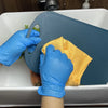 A person wearing blue nitrile gloves is using a yellow sponge to clean inside a white sink basin.