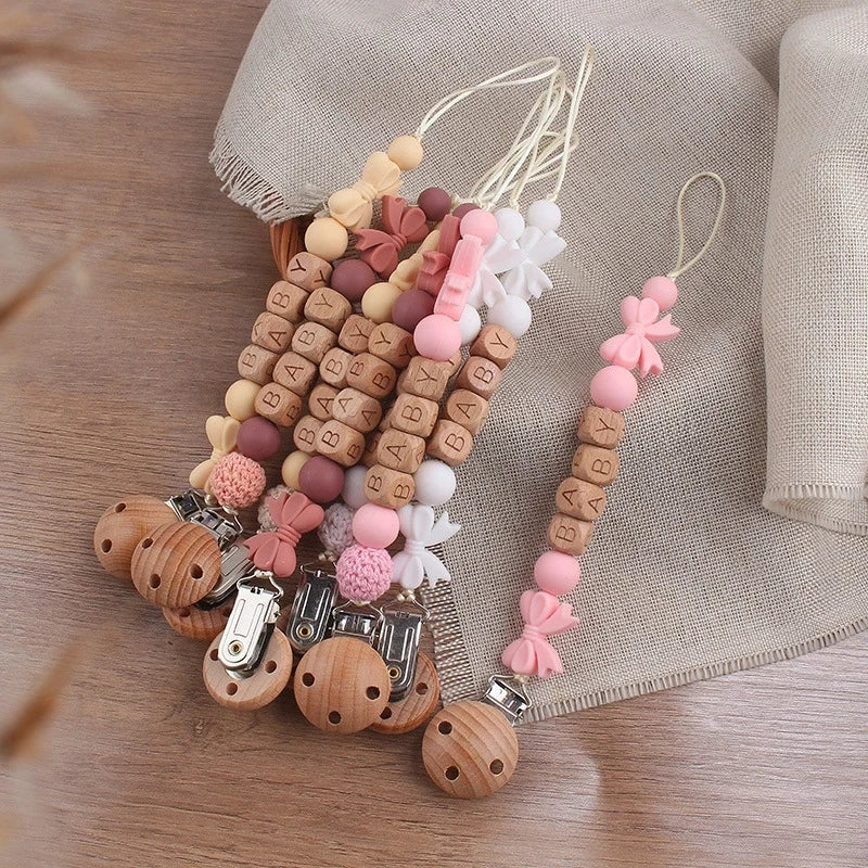 A collection of personalized silicone pacifier clips with various colors and names on a string against a beige cloth.