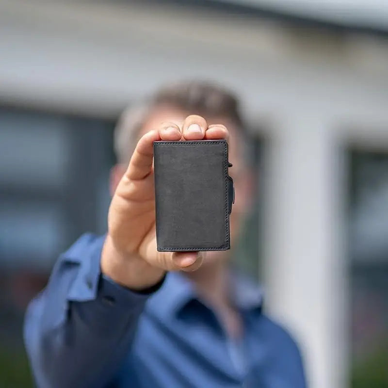A hand holds a dark gray RFID-blocking pop-up leather card holder wallet against a blurred background.