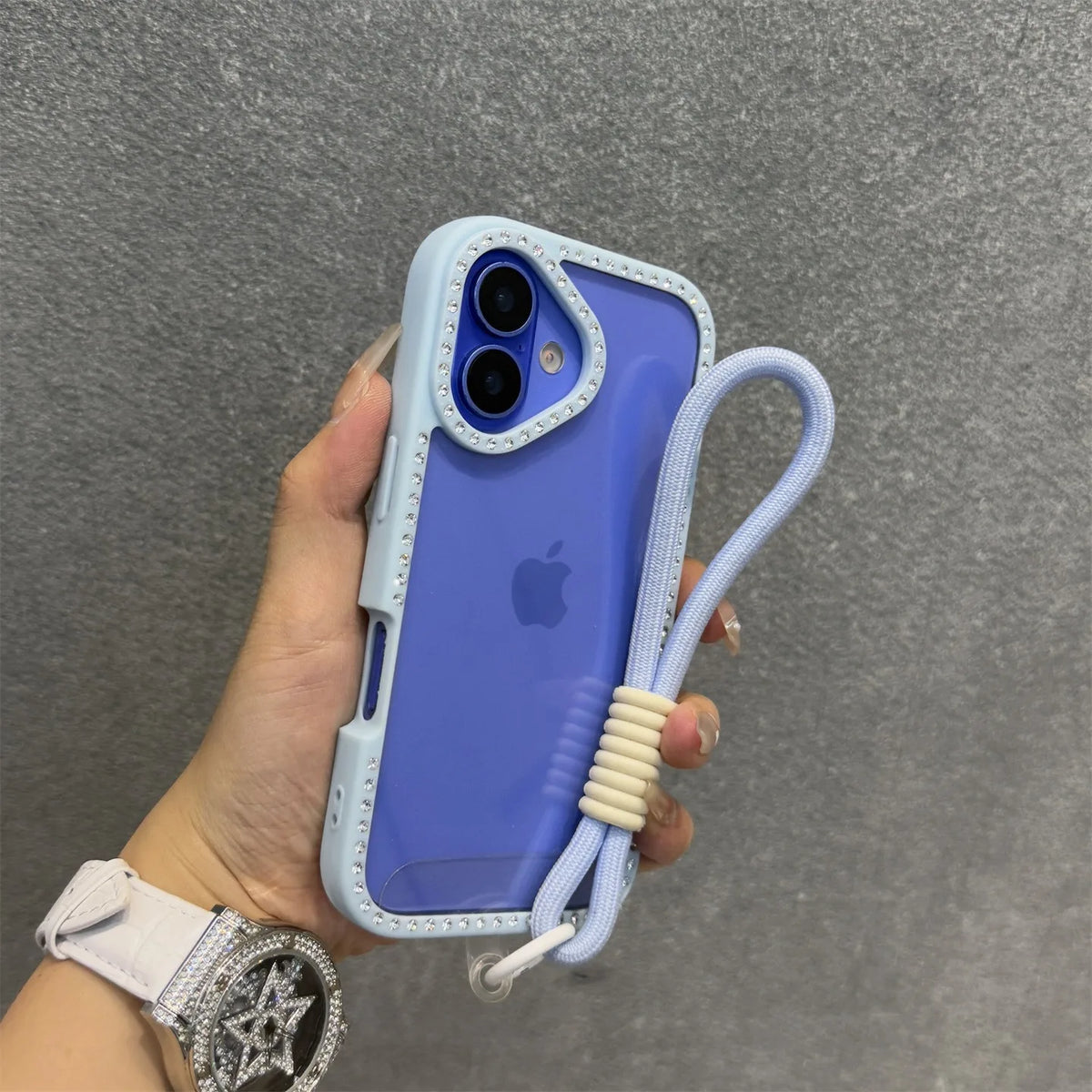 Una mano sostiene un iPhone azul claro en una funda transparente con correa blanca, contra un fondo gris.