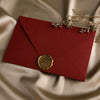 A deep red envelope with a gold sealing wax sticker and delicate dried floral sprigs resting on a silky fabric background.