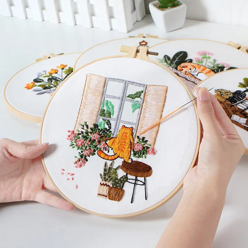 A hand is holding a round embroidery hoop featuring a detailed orange cat and floral designs on white fabric.