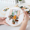 A hand is holding a round embroidery hoop featuring a detailed orange cat and floral designs on white fabric.