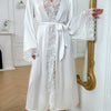 A white silk satin robe with intricate lace trim tied at the waist, displayed next to a gold-framed mirror.