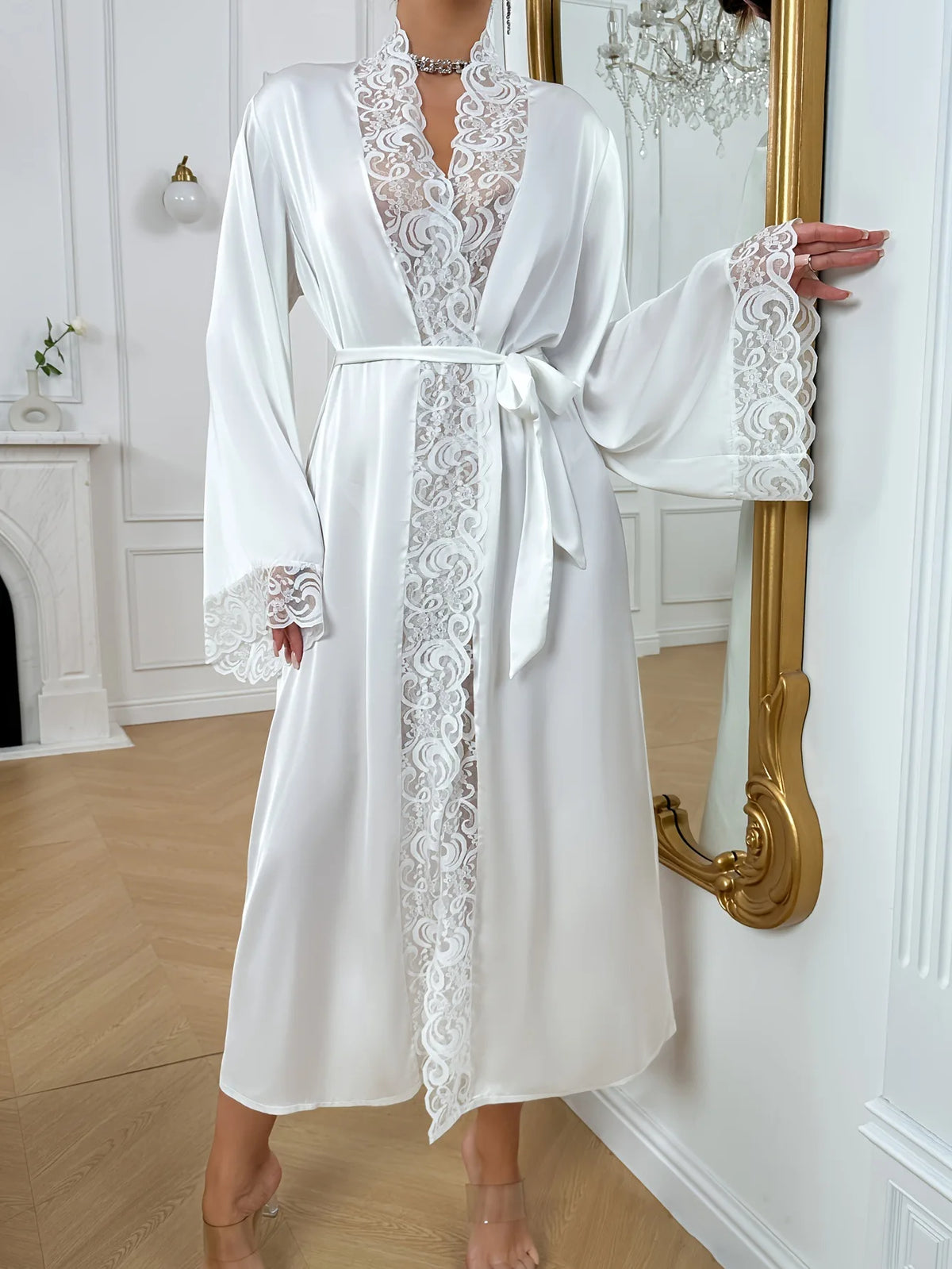 A white silk satin robe with intricate lace trim tied at the waist, displayed next to a gold-framed mirror.