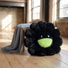 A black sunflower plush cushion with a smiling green face rests on a wooden floor next to a gray fabric.