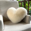 A fluffy, heart-shaped cream-colored faux fur throw pillow resting on a light gray sofa.