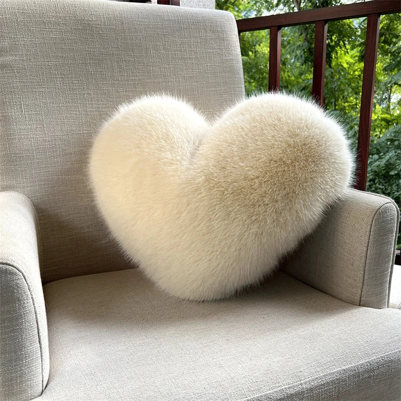 A fluffy, heart-shaped cream-colored faux fur throw pillow resting on a light gray sofa.