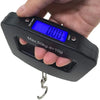 A hand holds a black portable digital luggage scale with a blue backlit display showing 0.00 and a metal hook.
