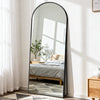 A full length oval mirror with a black frame reflecting a bedroom interior and sunlight.