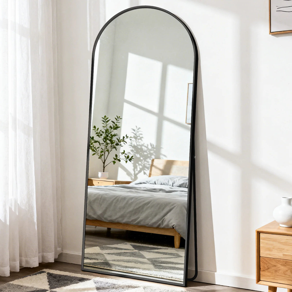 A full length oval mirror with a black frame reflecting a bedroom interior and sunlight.