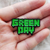 A bright green enamel pin shaped like the text GREEN DAY with a crossed guitar symbol, held in a person's hand.