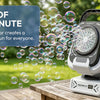 A white bubble machine is shown on a wooden table, creating a dense cloud of bubbles in a green outdoor setting.