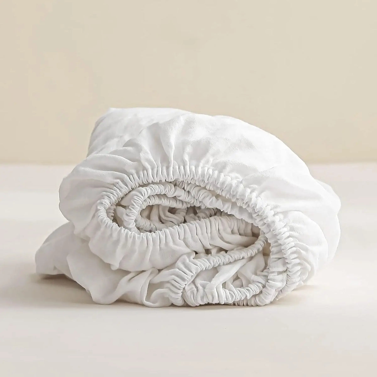 A rolled white linen fitted sheet with deep pockets is shown on a light surface.