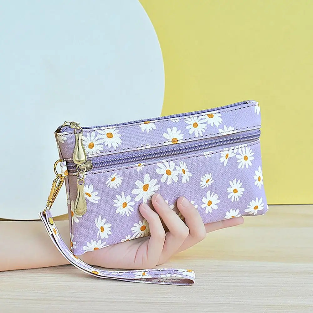 A hand holds a purple daisy print PU leather wristlet wallet with a zipper closure against a yellow background.