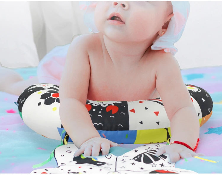 A baby is lying on a colorful mat, holding a high contrast tummy time pillow.