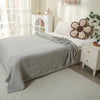 A light gray quilted blanket draped over a bed, next to a white nightstand and potted greenery.