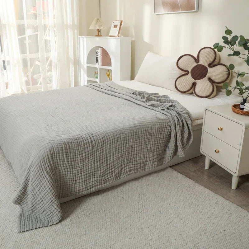 A light gray quilted blanket draped over a bed, next to a white nightstand and potted greenery.