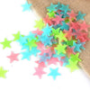 A pile of 100 luminous star-shaped wall stickers in various pastel colors like pink, blue, and green on a burlap background.