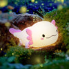 A glowing pink axolotl night light rests on mossy rocks in a dark, natural setting.