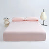 A pink cooling ice-silk fitted sheet with two pillows is displayed on a white surface next to a light fixture.